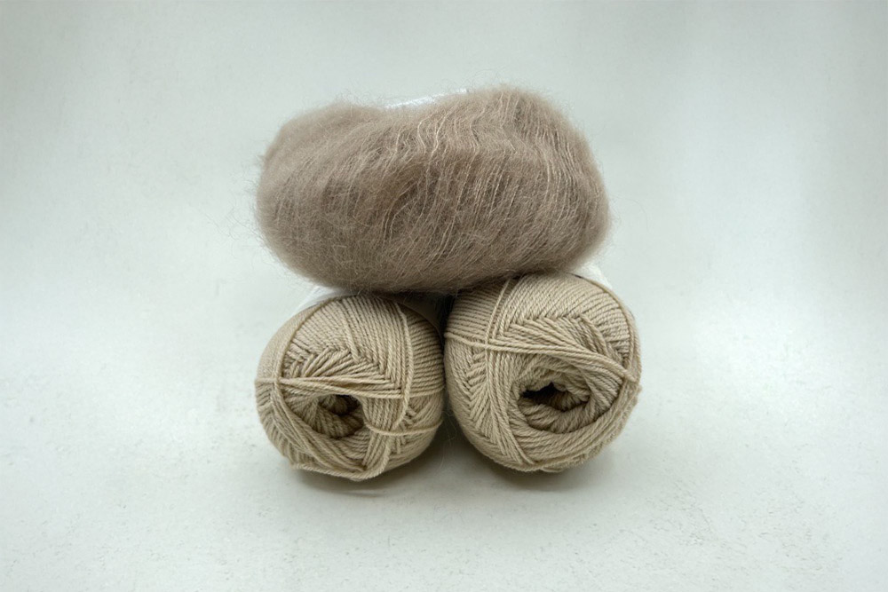 Tre nystan av beige garn på en vit bakgrund. Två nystan av slätt garn ligger underst och ett fluffigt mohairliknande nystan vilar ovanpå dem.