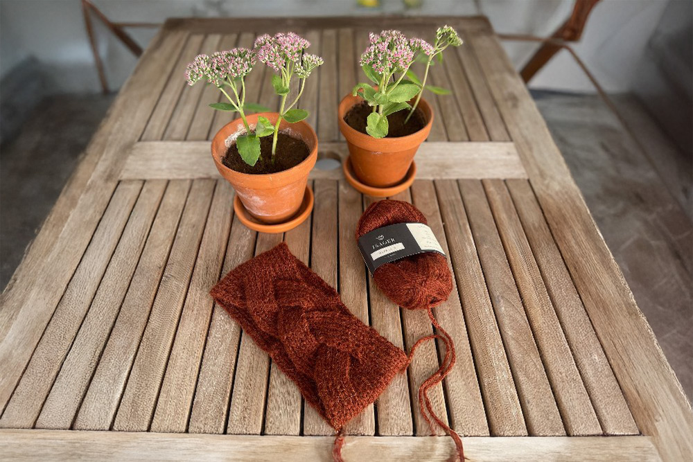 Garnpaket för stickat, brunt flätstickat pannband och ett nystan rödbrunt garn på ett träbord bredvid två krukväxter.